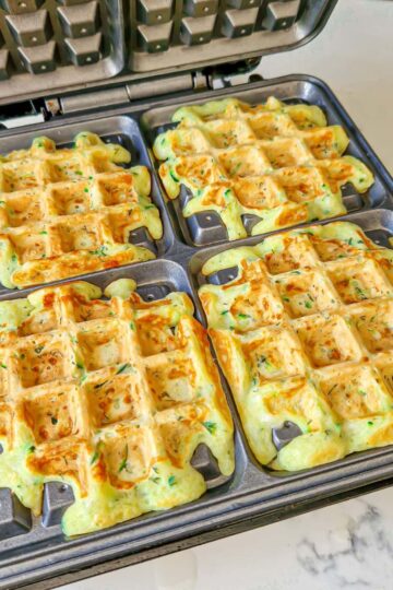 Four golden zucchini waffles cooking evenly inside a waffle maker.