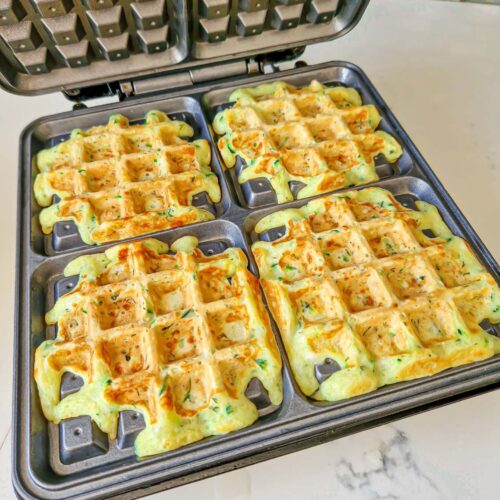 Four golden zucchini waffles cooking evenly inside a waffle maker.