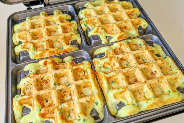 Four golden zucchini waffles cooking evenly inside a waffle maker.