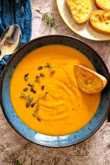 Creamy homemade pumpkin soup served in a bowl, topped with roasted pumpkin seeds and a drizzle of olive oil, with toasted bread in the background.