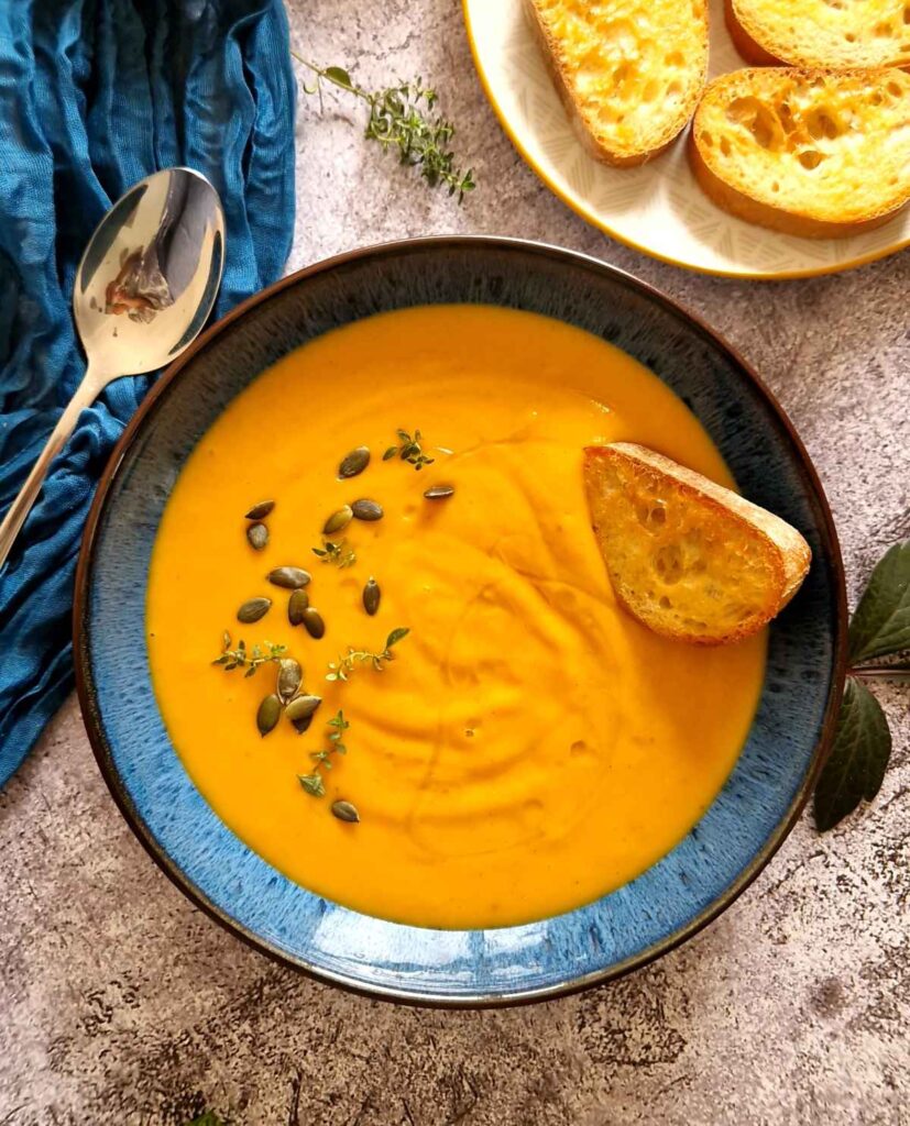 Creamy homemade pumpkin soup served in a bowl, topped with roasted pumpkin seeds and a drizzle of olive oil, with toasted bread in the background.