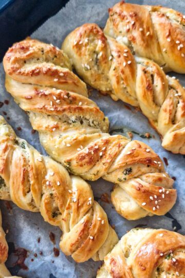 Golden-brown homemade garlic, cheese, and herb twists on a baking tray, topped with sesame seeds, freshly baked and ready to serve as a snack or appetizer.